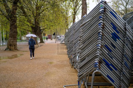 Téléchargez les photos : LONDRES - 24 avril 2023 : Une femme avec un parapluie et un sac à dos passe devant des barrières métalliques empilées haut sur le centre commercial, prête pour le couronnement du roi Charles. - en image libre de droit