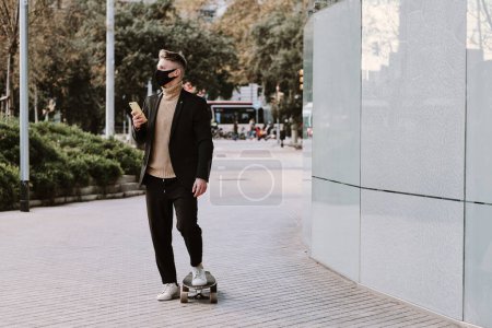 Téléchargez les photos : Homme avec masque facial protecteur utilisant un mobile en attendant dans la rue avec une planche à roulettes - en image libre de droit