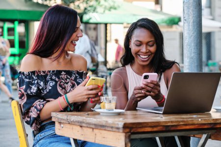 Foto de Dos mujeres multiétnicas felices usando móviles y un portátil mientras están sentadas en una cafetería - Imagen libre de derechos