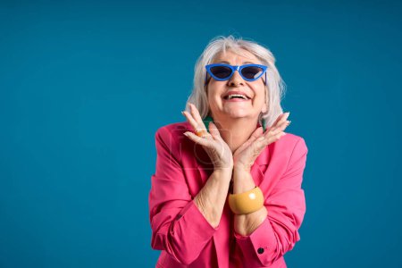 Fröhliche Seniorin mit schockiertem Gesichtsausdruck trägt blaue Sonnenbrille