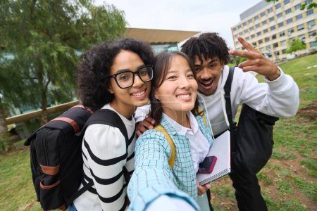 Three young people are smiling and posing for a picture in a park. One of them is wearing glasses and has a backpack on. The other two are also wearing backpacks. Scene is happy and friendly