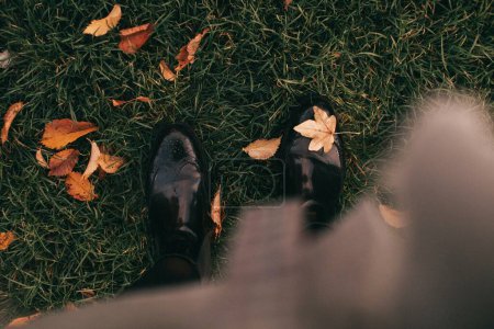 Woman in black boots stands on lawn grass with autumn leaves close upper view. Fall season fashion