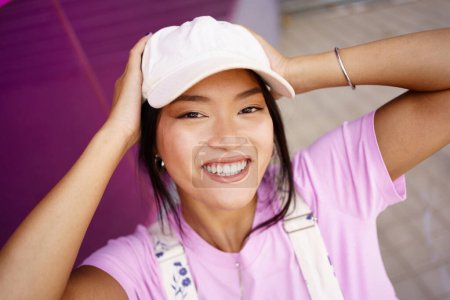 Téléchargez les photos : D'en haut heureux jeune asiatique femelle dans élégant chapeau toucher la tête et en regardant la caméra avec sourire sur la rue de la ville - en image libre de droit