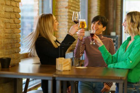 Foto de Las mujeres optimistas en ropa casual tintineo vasos de bebidas alcohólicas y proponer tostadas, mientras que de pie cerca de la mesa y la ventana en el día de fin de semana en el pub - Imagen libre de derechos