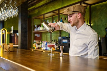 Téléchargez les photos : Homme barbu en chemise blanche et bouchon remplissage verre avec cocktail de shaker tout en se tenant derrière le comptoir et de travailler dans le bar - en image libre de droit