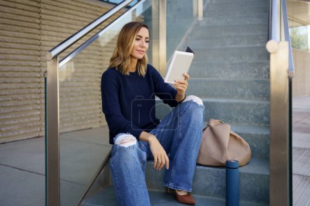 Foto de Mujer con ropa elegante sentada en las escaleras cerca de la bolsa y la lectura de libro electrónico en el día en la calle de la ciudad - Imagen libre de derechos