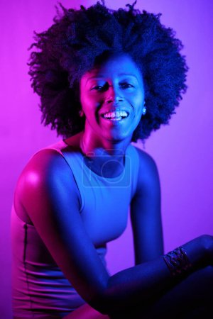 Foto de Feliz mujer afroamericana con el pelo rizado sonriendo y mirando a la cámara bajo la luz de neón violeta - Imagen libre de derechos