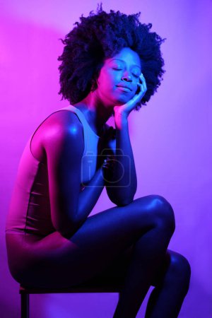 Téléchargez les photos : Vue latérale du modèle féminin afro-américain mince en body, avec les cheveux bouclés touchant l'épaule et regardant loin tout en étant assis sur des tabourets sous la lumière du néon violet - en image libre de droit
