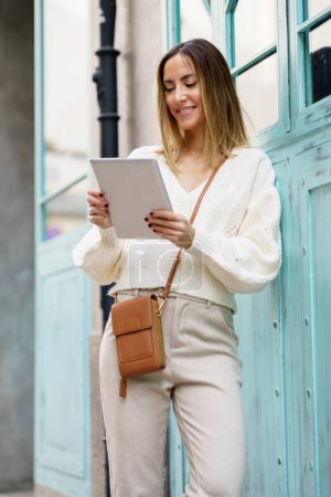 Foto de Mujer joven sonriente en ropa casual inteligente con bolsa marrón de pie en la calle de la ciudad mientras usa tableta que navega por las redes sociales - Imagen libre de derechos