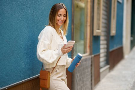 Téléchargez les photos : Joyeux jeune femme en tenue décontractée intelligente debout près du mur bleu tout en naviguant sur le téléphone mobile et la messagerie - en image libre de droit