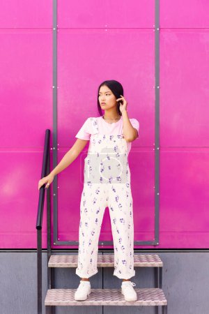 Téléchargez les photos : Jeune femme chinoise avec regard vide et expression sérieuse debout contre le mur rose du bâtiment moderne. Asiatique femelle en casual vêtements. - en image libre de droit