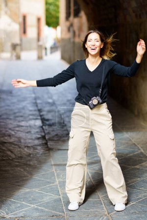 Photo for Full body of excited young female with long brown hair in casual clothes, smiling happily while standing in archway on old city street with photo camera during sightseeing trip - Royalty Free Image