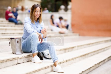 Foto de Mujer alegre de cuerpo completo en ropa casual sonriendo y navegando por las redes sociales en el teléfono celular mientras está sentada en las escaleras cerca de la bolsa durante el día en la calle de la ciudad - Imagen libre de derechos