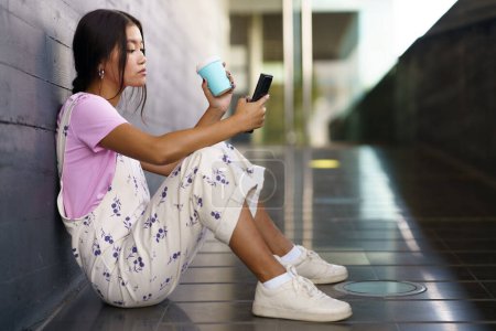 Téléchargez les photos : Full body young Asian woman in stylish clothes with cup of coffee to go siting on slope outside modern building and browsing smartphone in daytime - en image libre de droit
