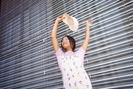 Téléchargez les photos : From below happy young Asian female in stylish clothes, smiling with closed eyes and raising arms with cap while dancing against metal wall on city street - en image libre de droit