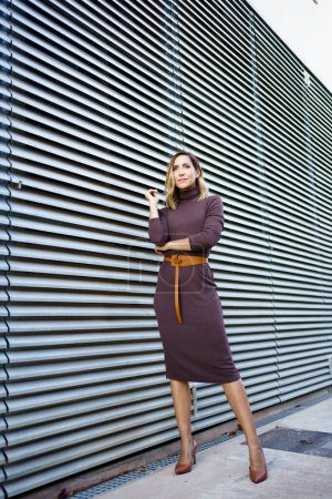 Téléchargez les photos : Full body self assured businesswoman in stylish dress with belt and high heeled shoes looking away while standing outside modern building on city street - en image libre de droit