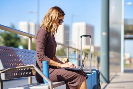 Téléchargez les photos : Female manager in stylish dress and sunglasses sitting on bench near suitcase and thermos and browsing netbook on station during business trip in city - en image libre de droit