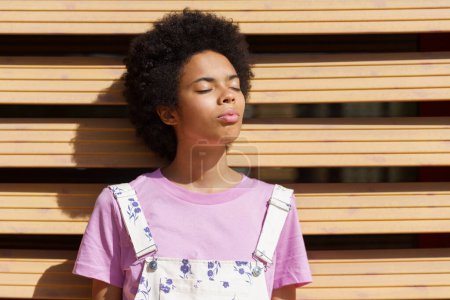 Téléchargez les photos : Charming African American female in casual wear standing under bright sunlight with closed eyes near wooden wall on street on summer day - en image libre de droit