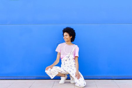 Photo for Full body of cheerful African American female in overall looking away with smile while sitting on haunches against blue background - Royalty Free Image