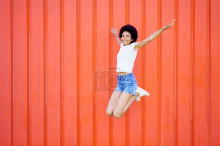 Téléchargez les photos : Full body of smiling African American female in casual outfit looking at camera while jumping with outstretched arms and bent knees against red background - en image libre de droit