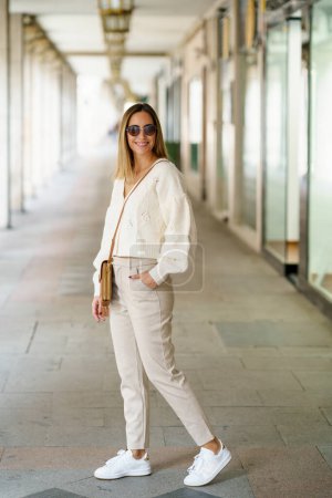 Full body of positive young female in beige blouse and pants with shoulder bag looking at camera smiling