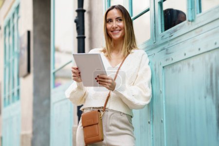 Téléchargez les photos : Cheerful blond haired female in smart casual clothes standing on city street while using tablet surfing internet - en image libre de droit