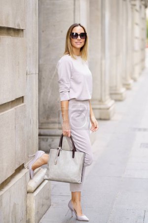 Photo for Full body of confident stylish woman gray outfit and sunglasses standing near old building with raised leg and holding handbag at city street - Royalty Free Image