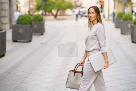 Téléchargez les photos : Side view of confident adult female manager in stylish outfit with blond hair looking at camera, while walking on paved street with laptop and bag in hands - en image libre de droit