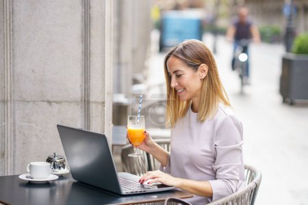 Foto de Side view of positive adult self employed lady with blond hair in casual clothes, drinking glass of fresh juice and smiling while working remotely on laptop in street cafe - Imagen libre de derechos