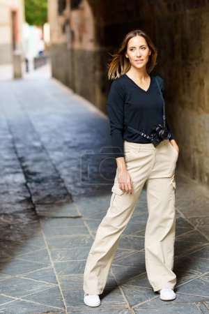 Photo for Full body of confident young woman with brown hair and photo camera, in casual clothes standing in stone archway with hand in pocket and looking away in old town - Royalty Free Image