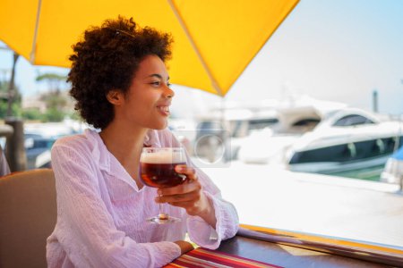 Téléchargez les photos : Cheerful young African American female in white blouse with curly hair smiling and looking away, while enjoying beer in street restaurant on sunny summer day on embankment - en image libre de droit
