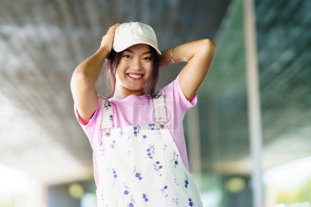 Téléchargez les photos : Merry young Asian woman in stylish t shirt and overalls with cap, looking at camera with smile and touching head on blurred background of contemporary building in city - en image libre de droit