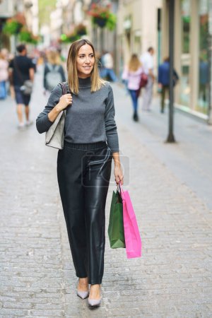 Téléchargez les photos : Full body of content adult woman in leather pants and heels carrying colorful paper bags after shopping and strolling on cobblestone town street smiling away - en image libre de droit