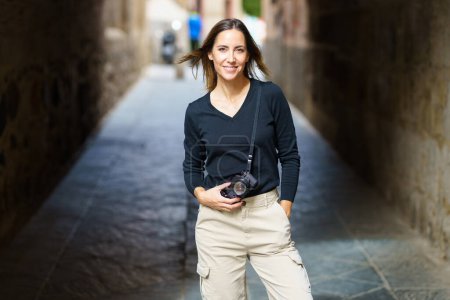 Téléchargez les photos : Self assured young female photographer with camera in casual clothes standing on narrow street with hand in pocket and looking at camera during vacation - en image libre de droit