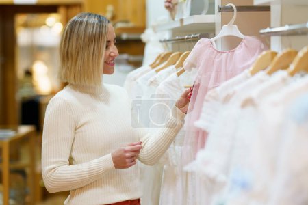 Foto de Positive female customer picking dress for girl while standing near rack with collection of stylish clothes in light modern boutique - Imagen libre de derechos