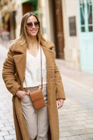 Foto de Confident female in sunglasses and brown coat with hand in pocket smiling while walking on town street - Imagen libre de derechos