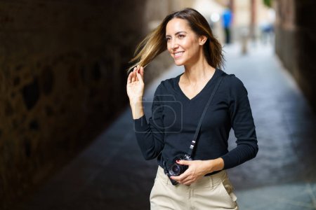 Foto de Content young female tourist in casual wear with retro photo camera, touching long brown hair and smiling while standing in narrow paved street in old town - Imagen libre de derechos