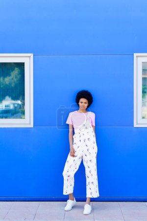 Téléchargez les photos : Full body of serious African American female model in casual wear looking away while standing near blue building in city - en image libre de droit