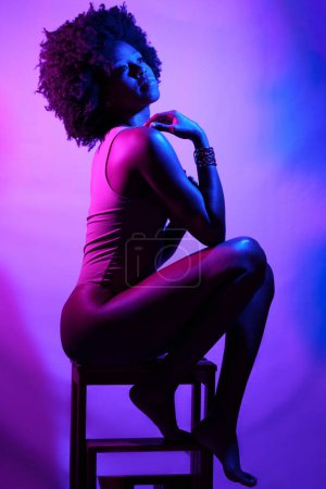 Foto de Vista lateral del delgado modelo femenino afroamericano en body, con el pelo rizado tocando el hombro y mirando hacia otro lado mientras está sentado en el taburete bajo la luz de neón violeta - Imagen libre de derechos