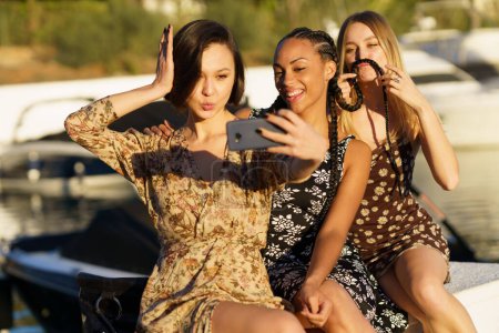 Foto de Delighted diverse female friends in dresses taking self portrait on smartphone while sitting together in wharf near river with boats - Imagen libre de derechos