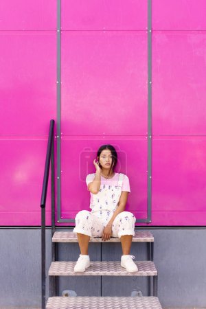 Foto de Cuerpo completo joven asiático hembra en ropa elegante tocando el pelo y mirando a la cámara mientras está sentado en las escaleras cerca de la pared rosa - Imagen libre de derechos