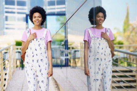 Photo for Happy African American young female with Afro hairstyle in overall looking away with smile while standing near glass building in city - Royalty Free Image