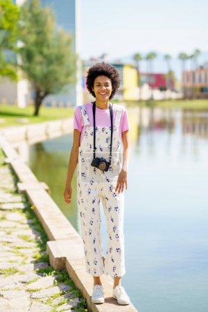Foto de Cuerpo completo de hembra afroamericana alegre con cámara fotográfica mirando hacia otro lado mientras está de pie cerca del lago en el soleado día de verano - Imagen libre de derechos