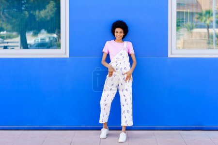 Photo for Full body of positive African American female in overall looking at camera while standing near blue house on paved street - Royalty Free Image