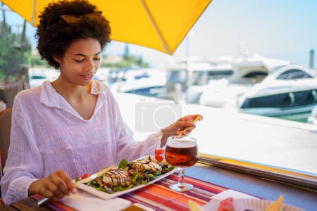 Foto de Feliz joven mujer negra en blusa blanca con el pelo rizado sonriendo y comiendo sabroso plato exquisito, mientras está sentado en la mesa sobre fondo borroso de terraplén en el soleado día de verano en el resort. - Imagen libre de derechos
