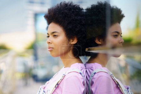 Photo for Side view of serious African American female with Afro hairstyle standing near glass house on street against blurred background in city - Royalty Free Image