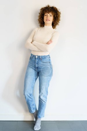 Téléchargez les photos : Full body of curly haired female in jeans and beige turtleneck leaning on white wall with hands crossed looking at camera - en image libre de droit