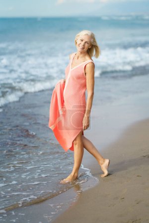 Foto de Hermosa mujer madura caminando a lo largo de la orilla de una playa tropical, vistiendo un bonito vestido naranja. Anciana disfrutando de su retiro en un retiro junto al mar. - Imagen libre de derechos