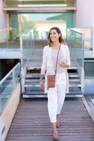 Photo for Full body happy adult woman in stylish clothes with smartphone smiling and looking away while walking on path against modern building - Royalty Free Image