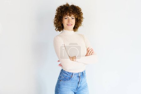 Foto de Confident adult curly haired female model wearing beige turtleneck and blue jeans standing with arms crossed against white background smiling at camera - Imagen libre de derechos